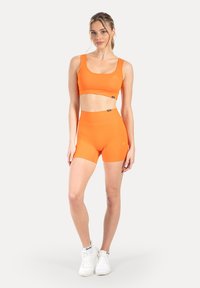 Fel oranje sportieve cropped top en shorts met hoge taille, gemaakt van gladde stof, met een subtiel logo detail. Witte sneakers maken de outfit compleet.