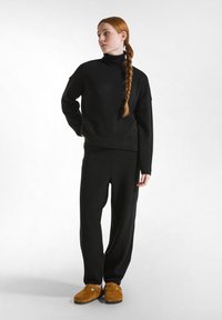 Schwarzer gestrickter Rollkragenpullover und passende, locker sitzende Hose, kombiniert mit braunen Wildleder-Slippern. Glatte Textur, minimalistisches Design.