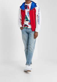 Röd och blå huva väst över en grafisk vit T-shirt, tillsammans med ljusblå jeans och vita sneakers. Funktioner inkluderar dragkedja och elastiska muddar.