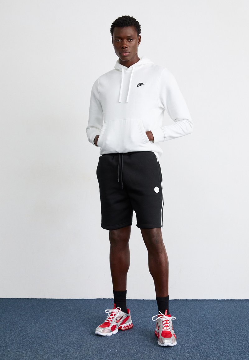 Hoodie Nike blanc avec cordons, shorts noirs avec bandes latérales blanches, et baskets Nike rouges avec accents gris. Debout sur un tapis bleu.
