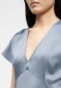 Γυναίκα που φοράει ανοιχτό μπλε σατέν φόρεμα με V-λαιμόκοψη και κοντά μανίκια, αποκαλύπτοντας το ντεκολτέ και το άνω μέρος του στήθους, πάνω σε απλό φόντο.