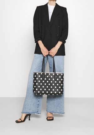 Sac fourre-tout noir à pois avec pois blancs, matériau texturé, doubles poignées et logo visible à l'avant. Le modèle porte un blazer noir et un jean.