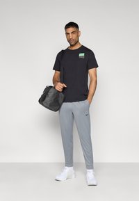 Μαύρο t-shirt με πολύχρωμο λογότυπο, γκρι φόρμα, λευκά αθλητικά παπούτσια και μαύρη τσάντα ώμου. Απλός, αθλητικός σχεδιασμός με χαλαρή εφαρμογή.