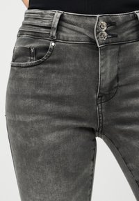 Κοντινό πλάνο γκρι τζιν ψηλόμεσου με διπλό μεταλλικό κουμπί κλεισίματος και λεπτομέρειες σε τσέπες μπροστά, φορεμένα με μαύρο τοπ.