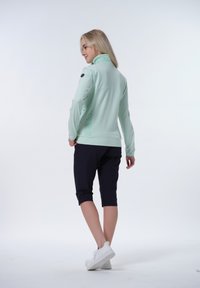 Ljust mintgrön zip-up jacka med hög krage, design med sidopanel och logodetalj på ärmen; kombinerad med svarta knälånga shorts.