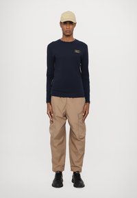 Jūrinė ilgomis rankovėmis marškiniai su auksiniu logotipu, derinami su chaki spalvos plačiomis kelnėmis ir juodais sportiniais batais. Kepurė yra smėlio spalvos, užbaigianti aprangą.