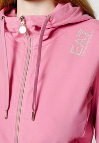 Rosa zip-up hoodie i bomullsblandning. Har hög krage, dragsko och metallisk EA-logotyp på vänster bröst. Slät textur.