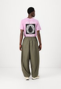 Lilla t-shirt med grafisk print på ryggen; parret med løse, olivengrønne bukser med sidelommer; neutrale sneakers er synlige.