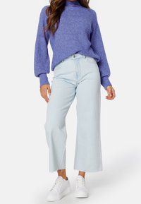 Jean bleu clair court avec ourlet effiloché, associé à un pull violet en maille avec un col montant et des manches bouffantes. Baskets blanches.