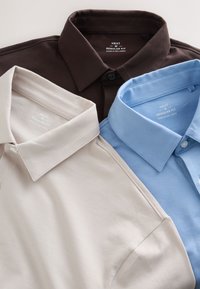 Trois polos de taille moyenne coupe régulière en marron, beige et bleu clair, marque « Next », fabriqués au Sri Lanka, soigneusement disposés.