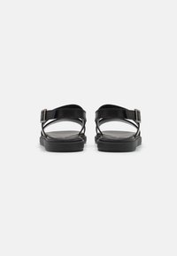 Sorte sandaler med et elegant design, kryssende reimer og spenne. Flat såle med en jevn tekstur og minimalistisk estetikk.