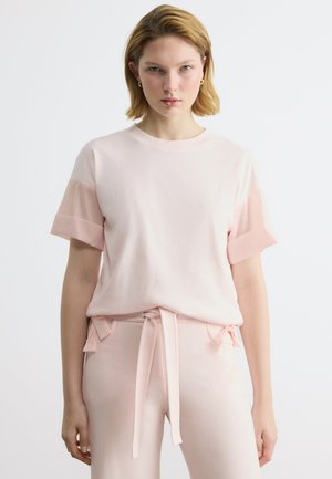Ung kvinde med kort blondt hår iført en lyserød kortærmet top og matchende bukser med et stofbælte, vendt fremad.