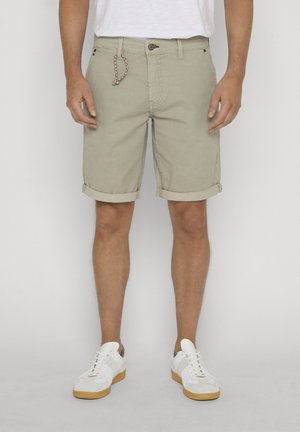 Mężczyzna w beżowych szortach o fakturze z podwiniętymi nogawkami, białej koszuli i białych trampkach z gumowymi podeszwami, stojący na jednolitym tle.