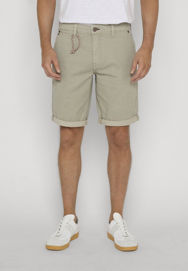 Homme portant un short beige texturé avec des revers roulés, une chemise blanche et des baskets blanches avec semelles en caoutchouc, debout devant un fond uni.
