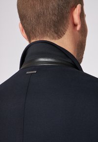 Cappotto navy con colletto alto con un accento in pelle e un piccolo etichetta metallica vicino al colletto, che mette in mostra un tessuto testurizzato simile alla lana.