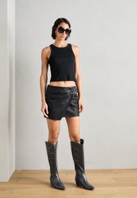 Top court noir sans manches, jupe portefeuille en cuir noir avec détail de boucle, et bottes noires montantes à bouts pointus. Textures lisses, design ajusté.
