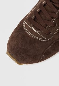 Detailní záběr na hnědou semišovou špičku tenisky s bílým prošíváním a hnědými tkaničkami na bílém pozadí.