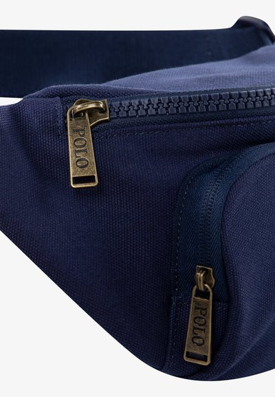 Marinfärgad tygfanny pack med dubbla dragkedjor i brons med "POLO"-logotyp, texturerad yta och rundade kanter. Kompakt design för mångsidighet.