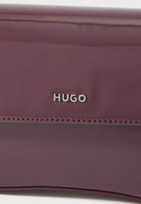 Peňaženka z platínovej lakovanej kože s prešitými detailmi a vyrazeným strieborným logom "HUGO". Úzky dizajn s hladkou textúrou a minimálnym kovovým príslušenstvom.