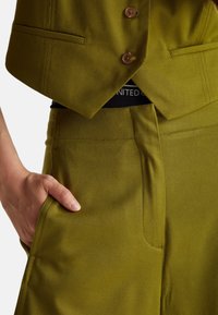Hand in de zak, gekleed in olijf groene op maat gemaakte broeken en een bijpassend geknoopt vest, waarbij de tailleband met gedeeltelijke tekst te zien is.