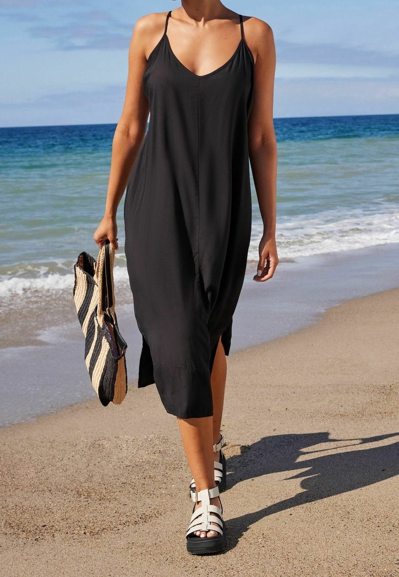 Femme marchant sur la plage portant une robe noire sans manches et des sandales blanches, tenant un sac cabas rayé avec des vagues de l’océan en arrière-plan.