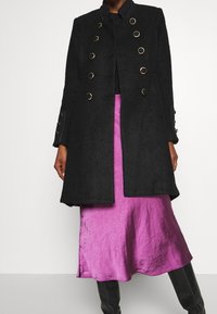 Abrigo de lana negro con botones circulares dorados, cuello alto y diseño ajustado; combinado con una falda midi brillante de color púrpura.