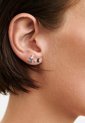 Lähikuva henkilön korvasta, jolla on lyhyet ruskeat hiukset ja hopeiset tähti- ja puolikuunappikorvakorut korvanlehdessä.