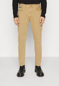 Beige puuvillachinot suoralla lahkeella, jossa on etutaskut, nappikiinnitys ja huomaamattomat ompeluyksityiskohdat. Mustat nilkkasaappaat jalassa.