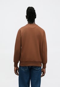 Sweat-shirt marron à col rond et poignets côtelés. Texture de tissu douce avec une couleur unie. Porté avec un jean bleu, vu de dos.