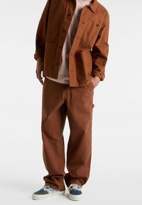 Rustfarvet jakke med mørke knapper og to forlommer, parret med matchende rustfarvede bukser. Grå og blå mønstrede sneakers fuldender looket.