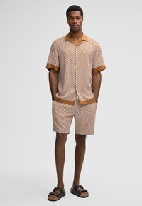 Man staat met zijn handen in zijn zakken, gekleed in een bijpassend bruin en wit gemusterd overhemd met korte mouwen en shorts, met zwarte sandalen.