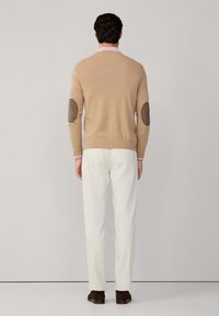 Uomo che indossa un maglione beige con toppe marroni sui gomiti, pantaloni bianchi, scarpe scure, in piedi con le spalle rivolte verso lo sfondo semplice.