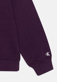 Manica di felpa di cotone viola con polsino a coste e logo "cK" ricamato in bianco. Tessuto liscio, design minimalista.