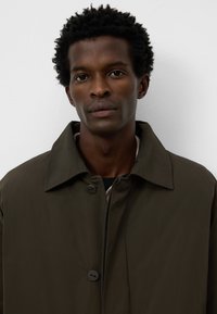 Cappotto leggero di un verde scuro, con colletto rialzato, due bottoni e un design pulito. Il materiale appare liscio con una leggera lucentezza.