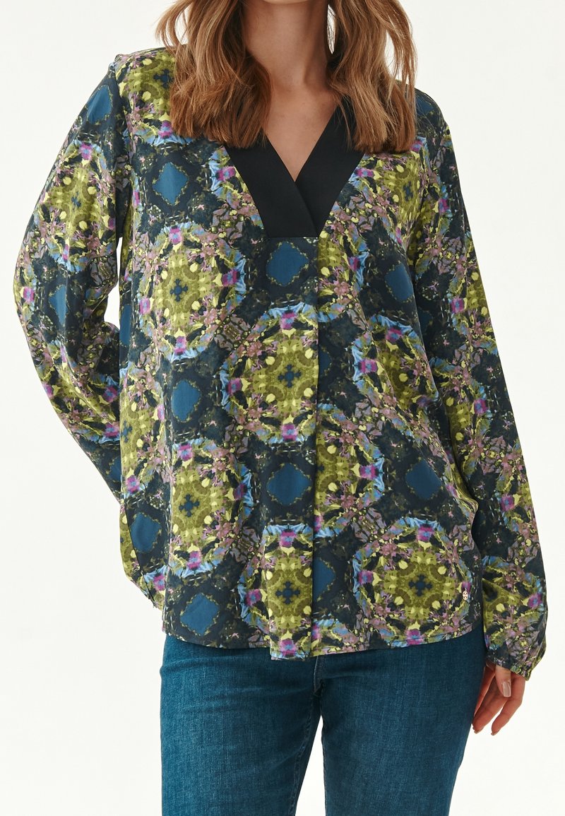 Femme portant un chemisier à manches longues avec un motif floral géométrique bleu, vert et rose et un col en V noir, assorti à un jean bleu.