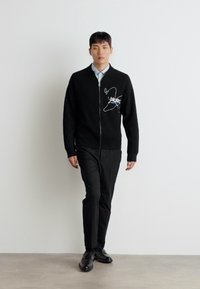 Svart ribbestrikket cardigan med glidelås, med hvit brodert design, båret over en lyseblå skjorte og paret med svarte bukser og sko.