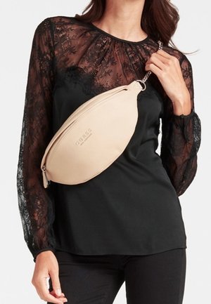 Kobieta ubrana w czarną koronkową bluzkę z długim rękawem i czarne spodnie, niosąca beżową torebkę GUESS na pasku łańcuszkowym przewieszoną przez klatkę piersiową.