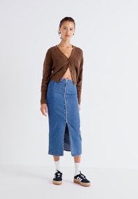 Rjav rebrast kardigan, denim midi krilo z sprednjim razporkom, bele nogavice in črne superge z svetlim podplatom. Enostaven in sproščen dizajn.