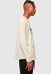 Hellbeige Langarm-T-Shirt mit lockerer Passform, ausgestattet mit schwarzem Drucktext und Akzenten, Rundhalsausschnitt und gerippten Bündchen.