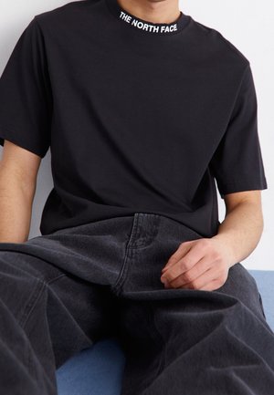 Schwarzes T-Shirt mit kurzen Ärmeln und weißem Logo am Kragen, kombiniert mit dunkelgrauer Denim-Jeans. Einfaches Design, weicher Stoff, lockere Passform.