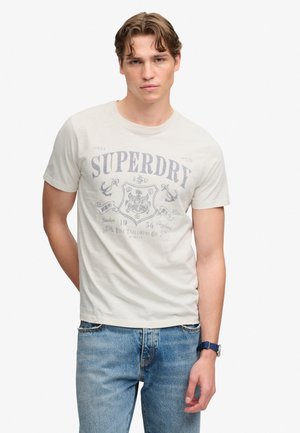 Junger Mann trägt ein hellbeiges kurzärmeliges T-Shirt mit verblasstem Superdry-Logo und blaue Jeans, steht mit einer Hand hinter dem Rücken.