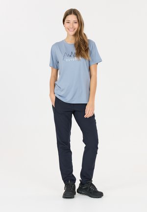 Junge Frau lächelt, trägt ein hellblaues T-Shirt mit Baummotiv und dem Wort "OUTDOOR", dunkle Hosen und schwarze Wanderschuhe, steht mit einer Hand in der Tasche.