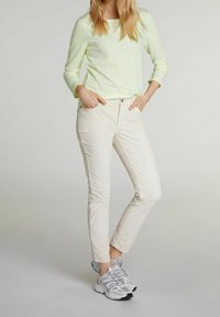 Žena ve světlém zeleném svetru s dlouhým rukávem, v bílých slim-fit kalhotách a bílých šedých sportovních botách, stojící s rukama v kapsách.