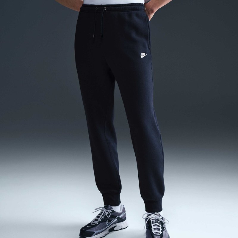 Persona che indossa pantaloni da tuta Nike neri e sneakers Nike nere e grigie, mani in tasca, in piedi contro uno sfondo scuro e uniforme.