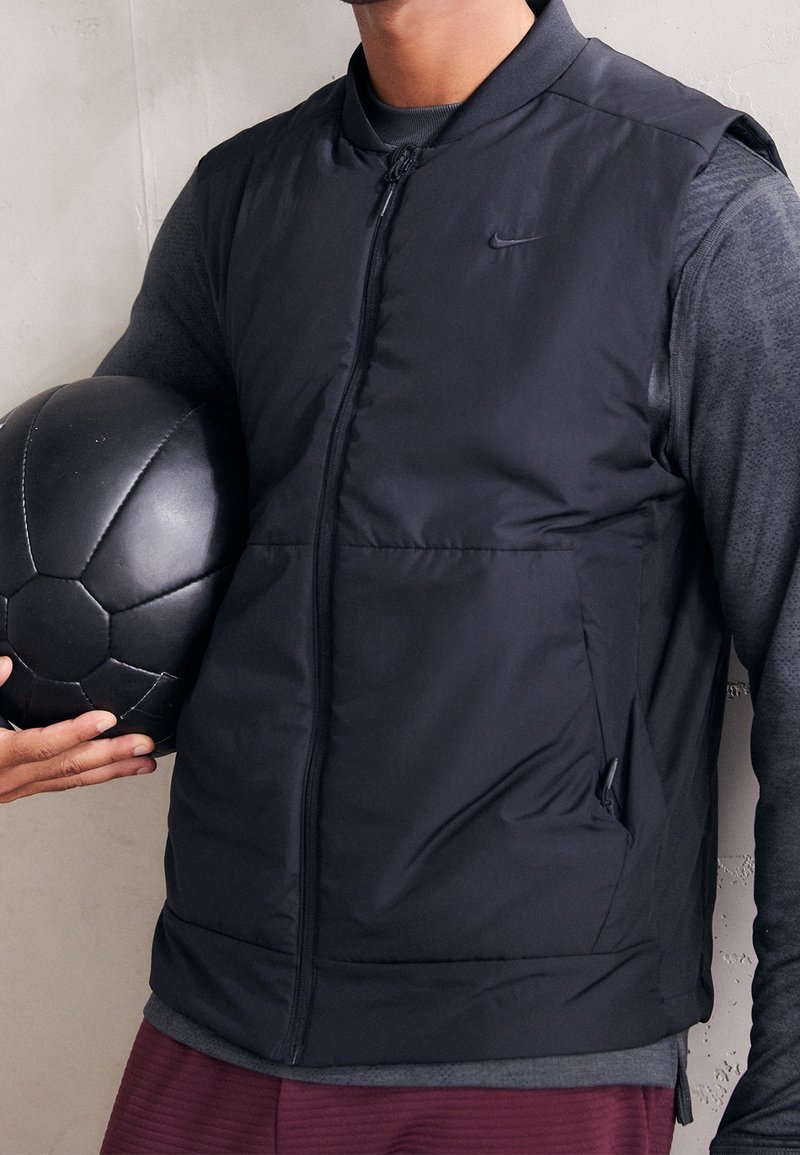 Schwarze gepolsterte Weste mit Reißverschluss, Seitentaschen und geripptem Kragen. Trägt man über einem dunklen Langarmshirt und hält einen schwarzen Fußball.