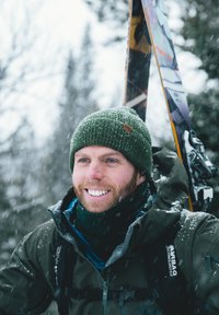 Snowy landscape with a man wearing a green knit beanie, dark jacket, and scarf, holding skis. Snowflakes falling around him.