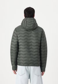 Homme portant une veste à capuche matelassée vert foncé et un jean bleu clair, de dos contre un fond blanc uni.