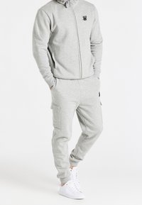 Felpa grigia con zip, cappuccio e logo, abbinata a pantaloni jogger coordinati con tasche cargo. Sneakers bianche completano il look.