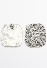 Silver metal pins with textured surfaces. One pin has a smooth, slightly curved shape and a central attachment. The other features a patterned design.