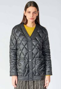 Veste matelassée noire avec col en V, fermeture à boutons, deux poches et accents texturés le long du col et de l'ourlet. Matière douce et brillante.